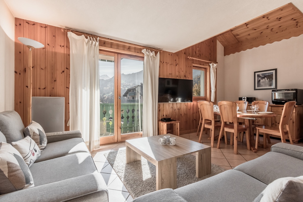 A spacious living area is showcased with light-coloured furniture and wooden accents. Large windows invite natural light and offer views of the surrounding mountains. A dining table for four and a cozy seating arrangement are visible, creating a welcoming atmosphere for relaxation.