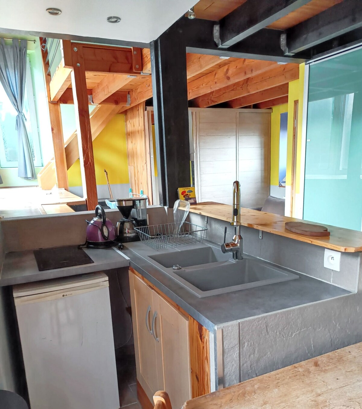 The kitchen area features a functional design with a stainless steel sink and wooden countertop. Equipped with essential appliances, including a kettle and refrigerator, it offers a space for meal preparation. Natural light enters through a nearby window, enhancing the overall brightness.
