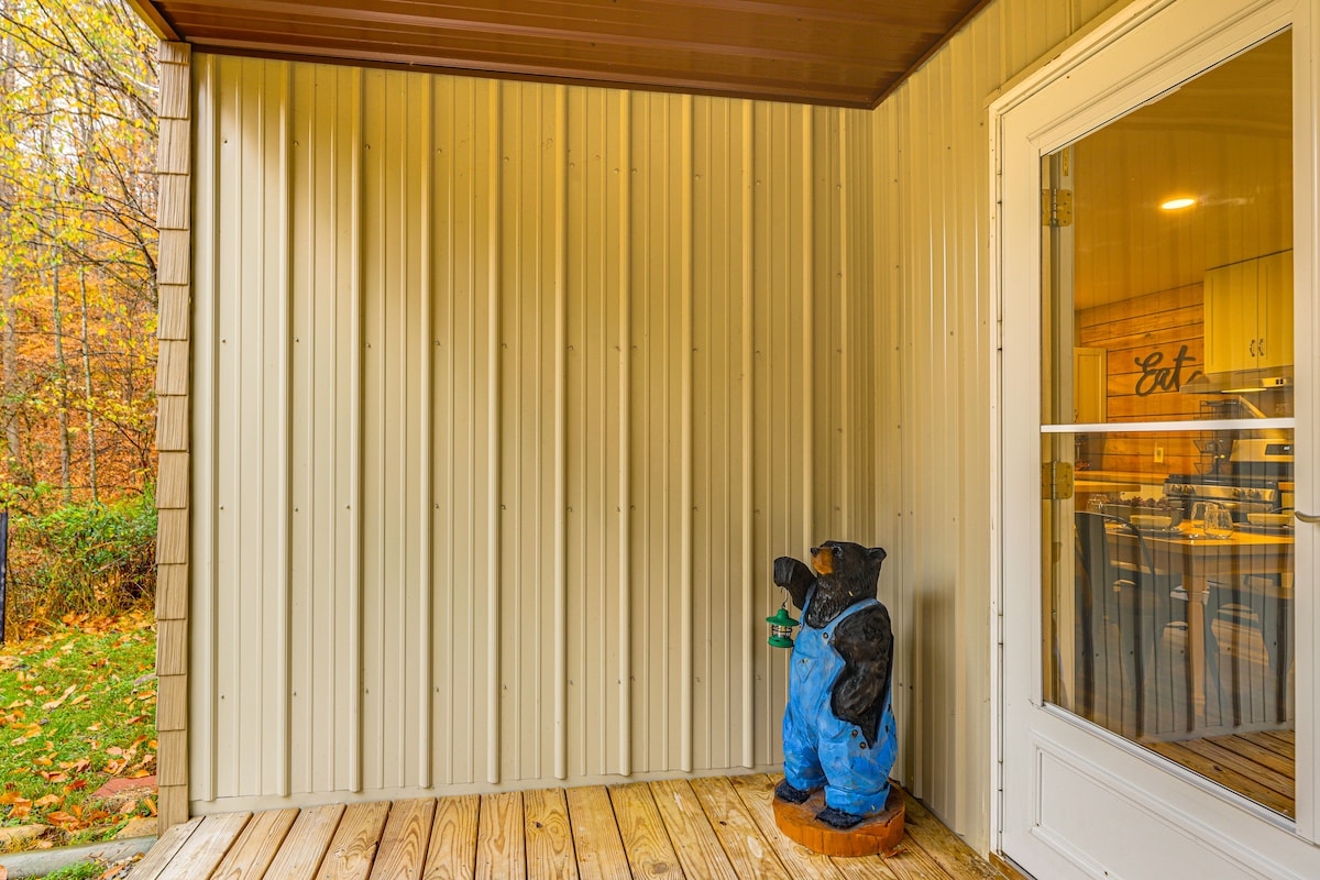 A front porch features a wooden floor and a wall of vertical metal siding. A bear statue dressed in blue overalls stands next to the entrance, adding a whimsical touch to the outdoor area.