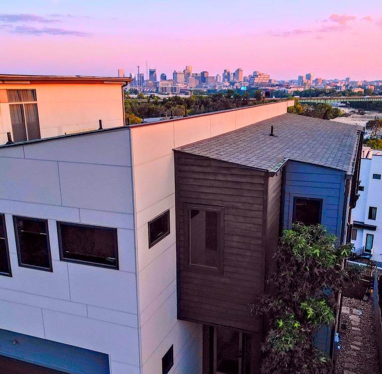 The exterior of the modern multi-story home features a mix of light and dark siding. A glimpse of the Nashville skyline is visible in the background, accompanied by a pastel sky at dusk. Nearby greenery contributes to the serene urban setting.