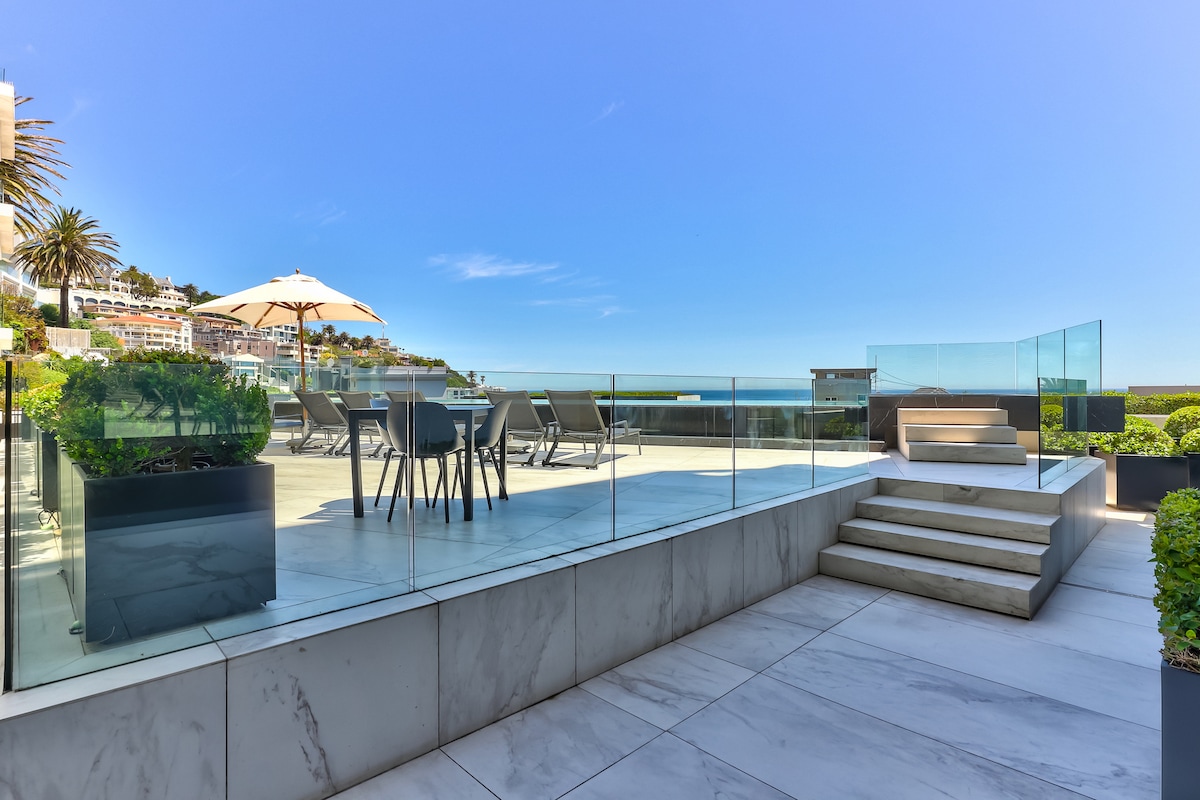 An outdoor terrace is showcased, featuring elegant glass railings that offer unobstructed views of the Atlantic Ocean. Sun loungers and a dining table for outdoor meals are visible, surrounded by lush greenery. Bright sunlight enhances the modern marble flooring and steps leading down to the lower area.
