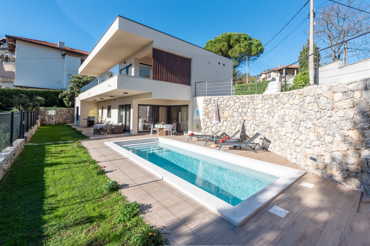 A modern villa is displayed, featuring a private outdoor pool surrounded by a wooden deck and neatly trimmed grass. Comfortable lounge chairs are positioned near the pool. The villa's contemporary architecture showcases large windows and a spacious patio area, providing a bright and inviting environment.