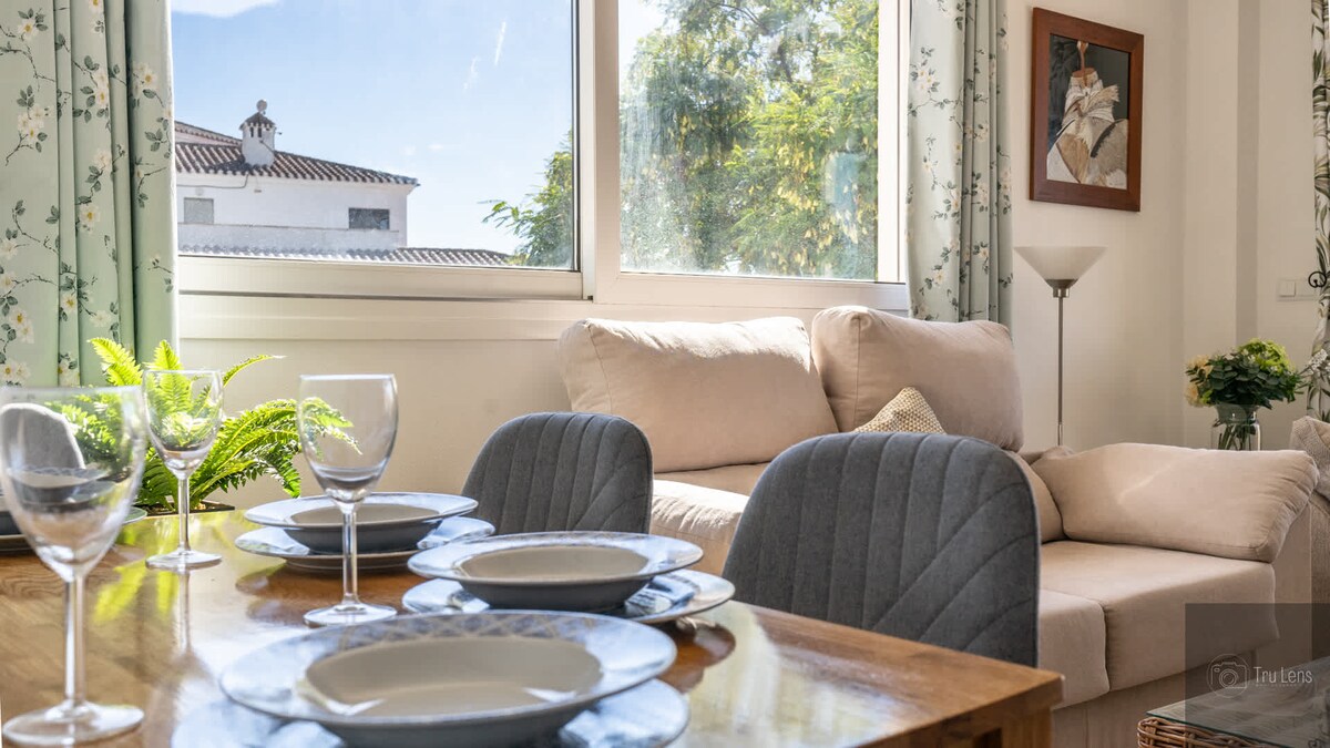 A dining table is set for four, featuring stylish plates and glassware. A seating area is highlighted by a light-colored sofa positioned near large windows, allowing natural light to brighten the space. Decorative plants and artwork add character to the room.