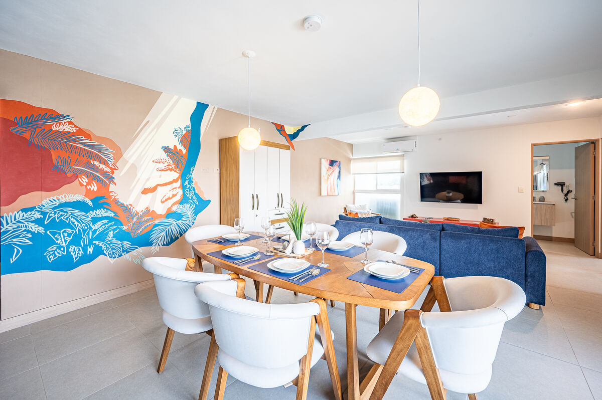 A dining area features a wooden table surrounded by six white chairs, set for a meal with plates and glasses. A mural of tropical foliage in vibrant colors decorates the wall. Soft lighting from pendant lamps creates a welcoming ambiance, with a view of the cozy living area in the background.