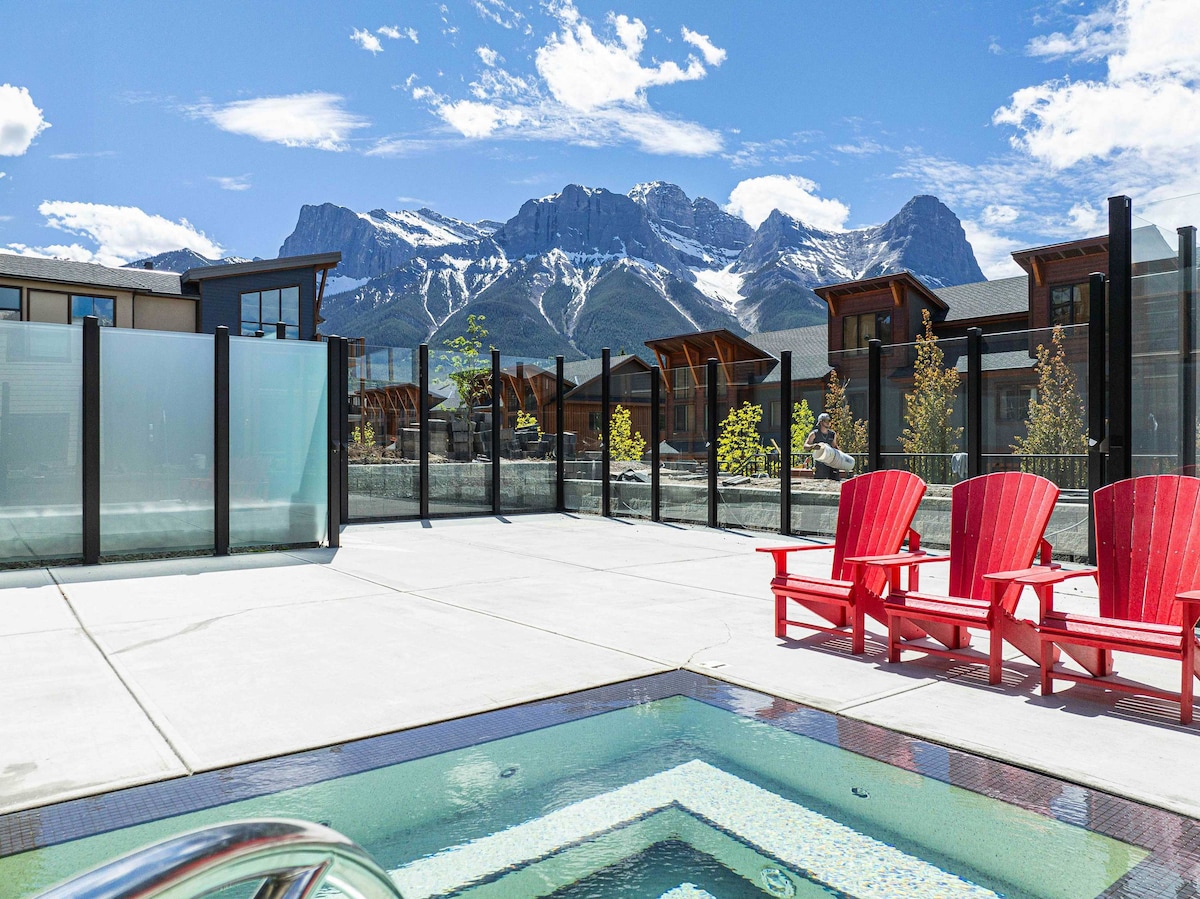 A shared outdoor hot tub is positioned on a spacious patio area, overlooking stunning mountain views. Two red Adirondack chairs are placed nearby on the concrete surface, complementing the serene landscape under a clear blue sky with scattered clouds.