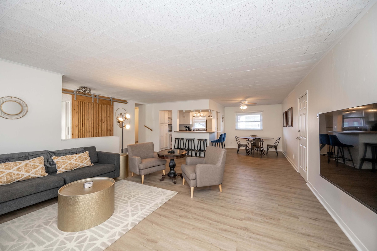 A spacious living room is furnished with a comfortable gray sofa and two armchairs positioned around a round coffee table. A sleek TV is mounted on the wall opposite, while a dining area with a circular table for four can be seen in the background. Natural light illuminates the entire space.