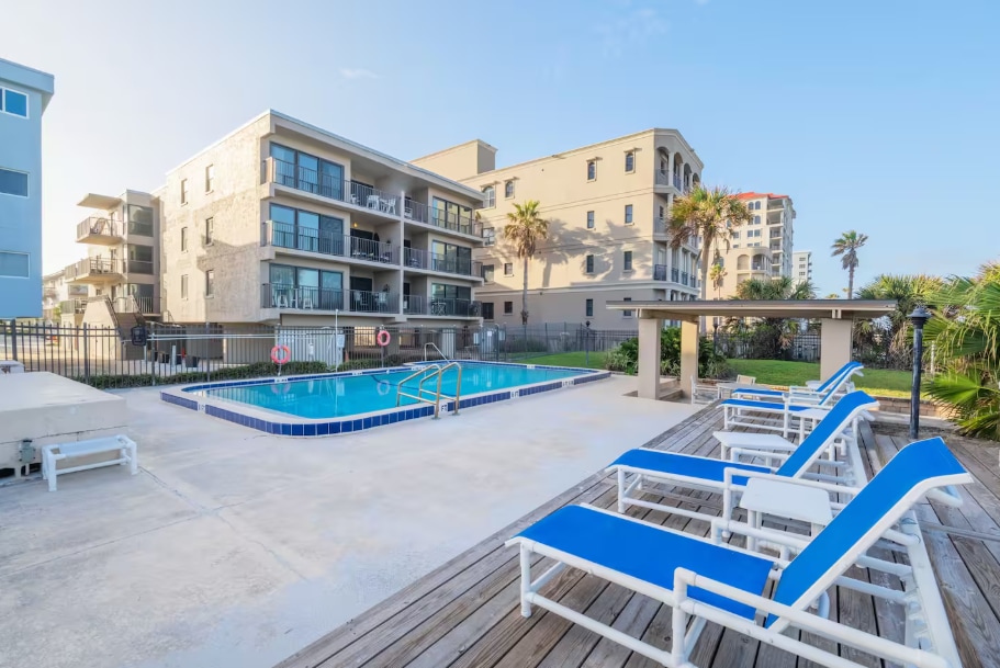 A community pool area features lounge chairs with blue cushions arranged around a sparkling water pool. The surrounding deck is made of weathered wood, with palm trees and coastal buildings visible in the background under a clear sky.