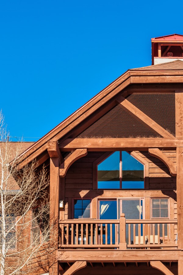 The exterior of a wooden vacation home is displayed, featuring a sloped roof and a prominent balcony with railings. Large windows capture natural light, reflecting a clear blue sky above. The surrounding trees are bare, indicating a seasonal transition.