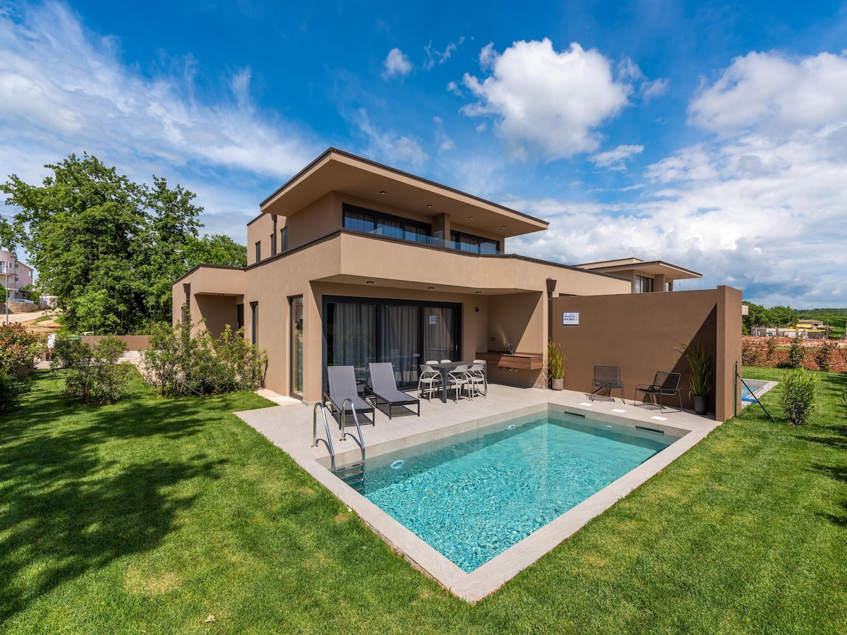 A modern luxury home is surrounded by lush greenery, showcasing a private swimming pool at the forefront. The spacious terrace features sleek lounge chairs and a welcoming seating area, while large windows reflect abundant natural light, enhancing the overall elegant design.