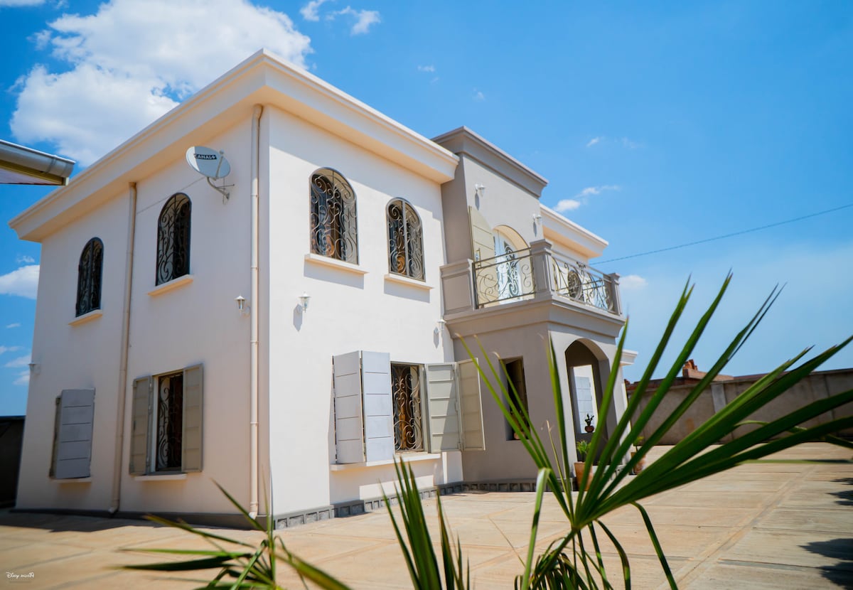 The villa features a modern two-story exterior with large windows and decorative metal grilles. A spacious balcony is visible on the upper level, enhancing the architectural appeal. Lush greenery surrounds the property, complementing the inviting outdoor space.