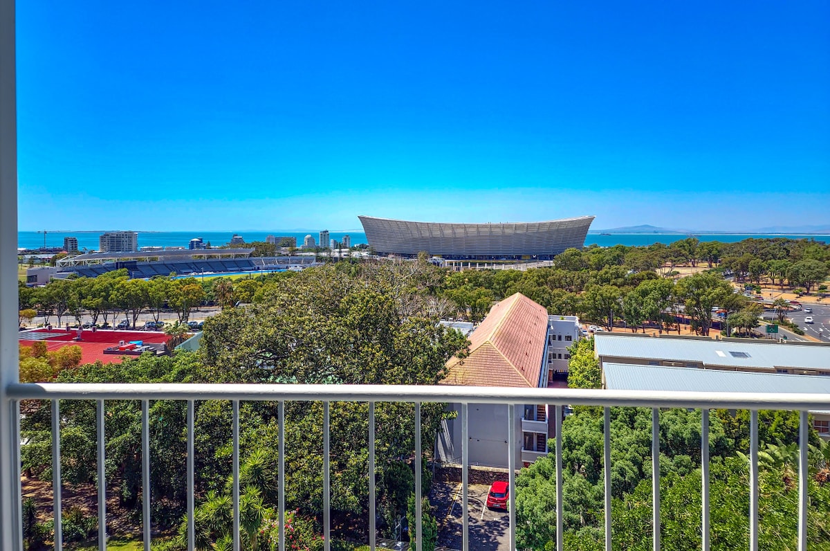 The image showcases a view from a balcony highlighting a large stadium and lush green trees below. The ocean can be seen in the distance under a clear blue sky, offering a bright and open perspective of the surrounding area.