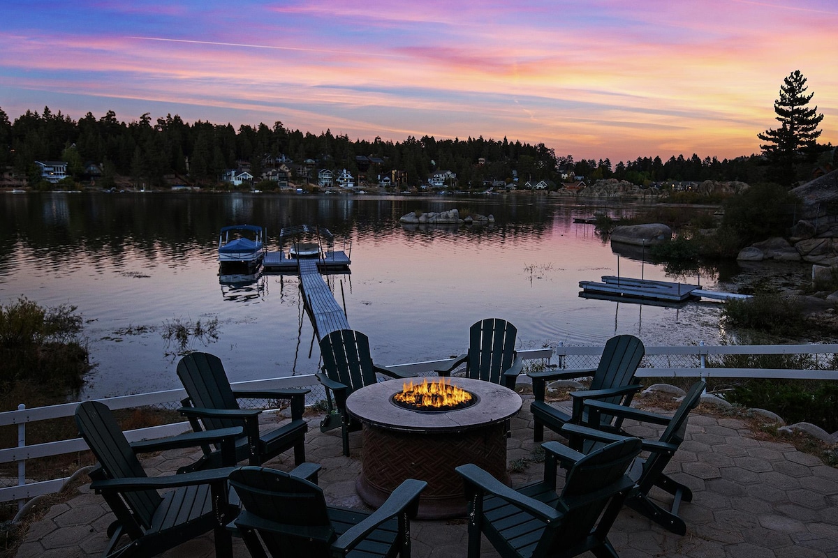 An outdoor fire pit surrounded by Adirondack chairs is set on a patio overlooking the lake, reflecting a vibrant sunset sky. A private dock is visible in the background, providing access to the serene water and natural landscape.