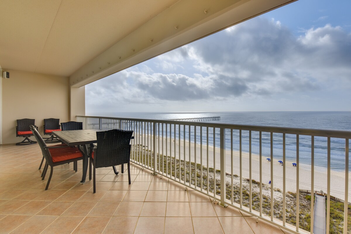 An oceanfront balcony offers a spacious outdoor space with a large dining table and black chairs, overlooking the beach and water. The view includes a distant fishing pier under partly cloudy skies, with soft waves gently lapping at the shore.