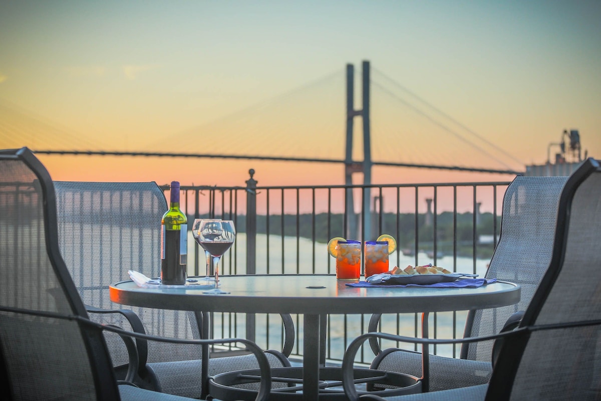 A round table set for two on the rooftop deck offers glasses of wine and colorful drinks, complemented by fresh snacks. The Savannah River is visible in the background, framed by a striking bridge and a soft sunset sky.
