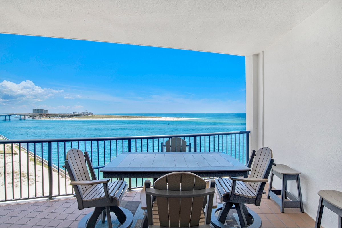 A spacious balcony is furnished with a rectangular dining table surrounded by six chairs, overlooking the Gulf waters. Panoramic views of the coastline, boat traffic, and distant shorelines are visible, complemented by a clear blue sky.