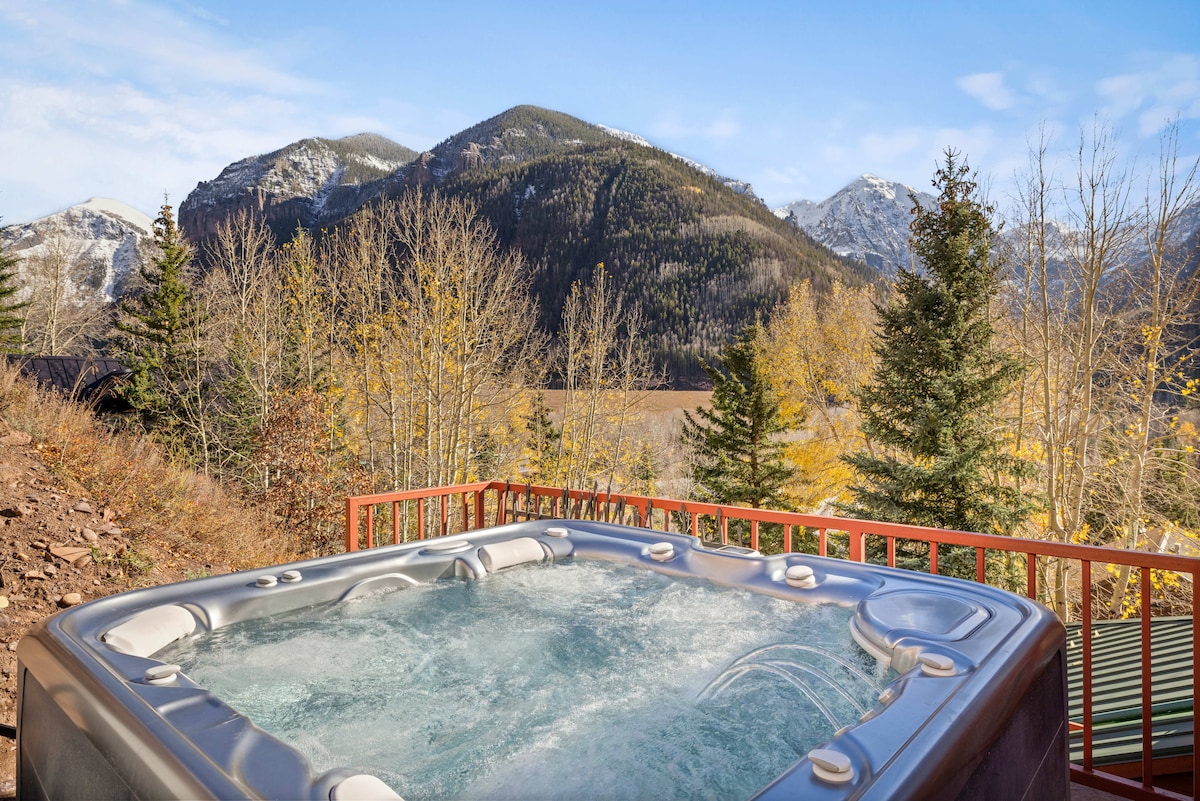 A hot tub is positioned on a wooden deck, surrounded by trees with autumn foliage. The expansive views encompass mountains in the distance, featuring varying shades of green and brown under a clear blue sky.