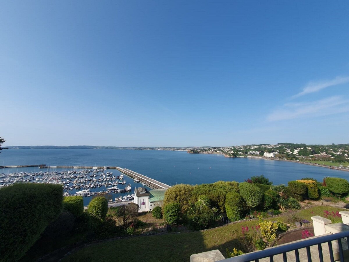 A panoramic view showcases the tranquil sea with numerous boats docked in the marina. Lush greenery borders the foreground, while gentle hills and coastal buildings can be seen in the distance, creating a serene atmosphere under a clear blue sky.
