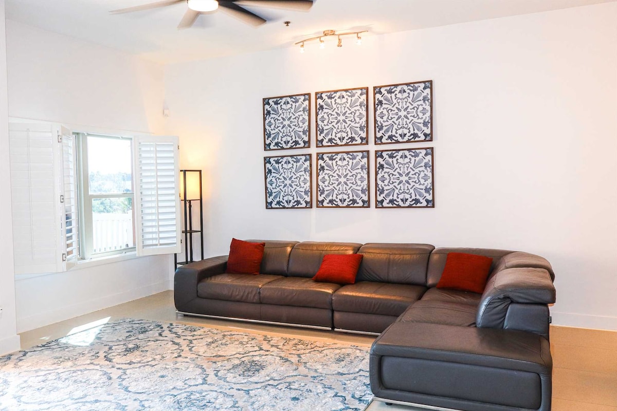 A spacious living area features a dark leather sectional sofa accented with red cushions. Four framed decorative panels hang above, contributing to the room's aesthetic. Natural light filters through the window, complemented by white plantation shutters, while a light-colored area rug adds texture underfoot.