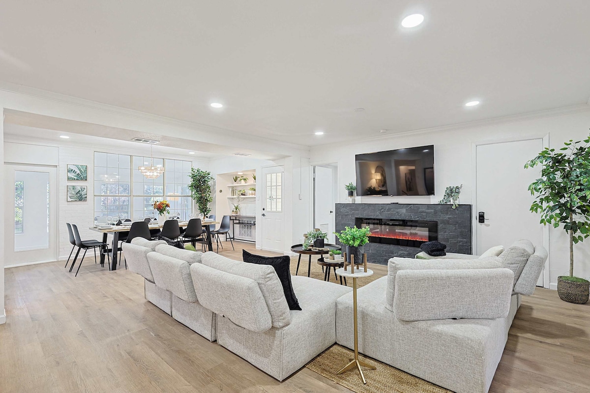 A modern living area features a spacious sectional sofa arranged around a sleek coffee table. A mounted television is situated above a contemporary fireplace, with light streaming through large windows. An elegant dining area with black chairs is visible in the background.