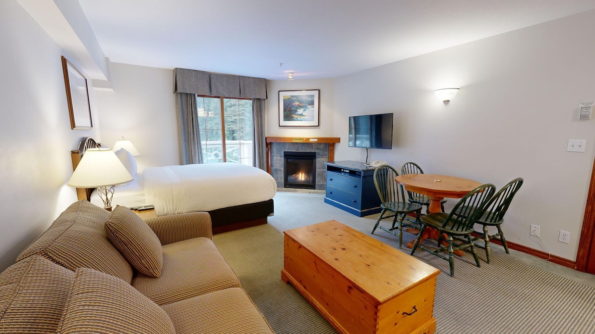 The studio features a comfortable layout with a spacious bed and a cozy sofa. A wooden coffee table sits in front of the sofa, and a dining table for four is present. Natural light enters through the window, enhancing the warm ambiance of the gas fireplace.
