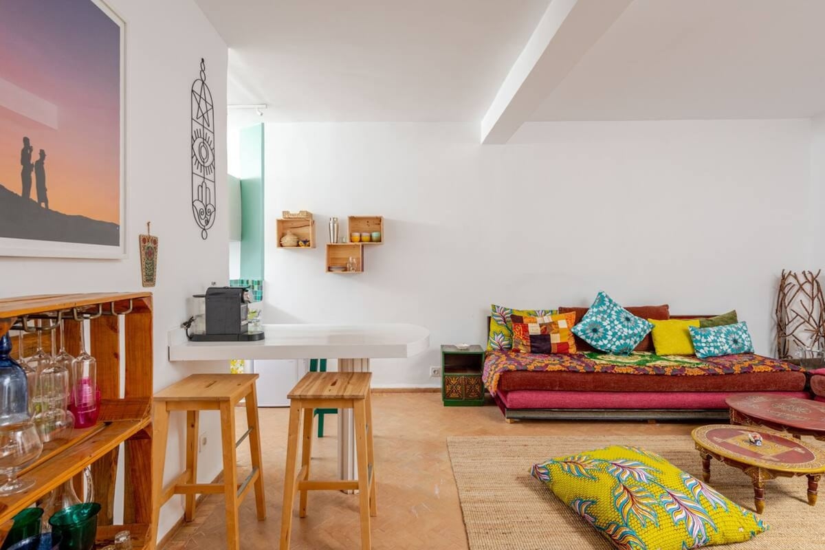 An inviting open-plan living area features a vibrant sectional sofa adorned with colorful cushions. A small dining table and stools stand nearby, alongside a wooden bar with glassware. Natural light fills the room, complementing the warm tones of the woven rug.