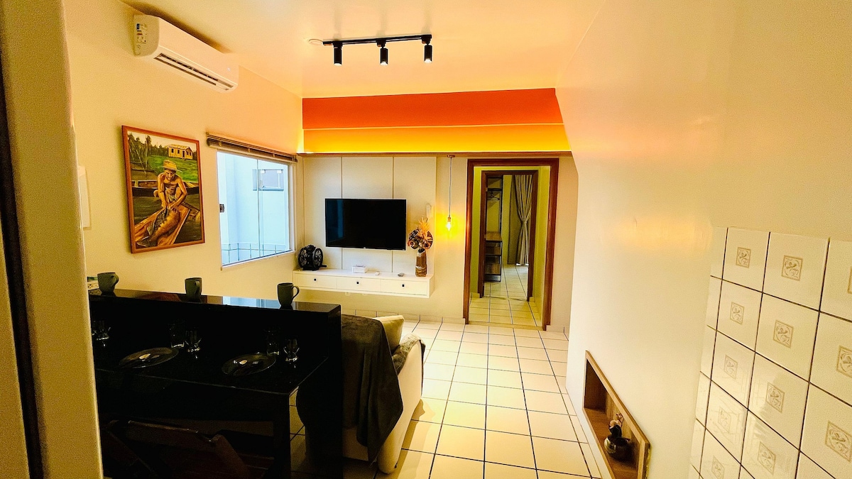 An open living space features a light-colored wall with an orange accent above a modern TV unit. A dining area with dark countertops is visible along with a comfortable sofa. Natural light enters through a window, enhancing the spacious feel of the room.