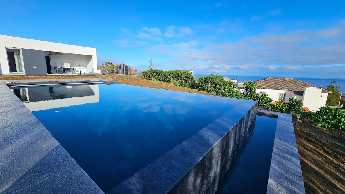 A private infinity pool reflects the clear blue sky, surrounded by a landscaped area with lush greenery. The tranquil water seamlessly merges with the horizon, offering views of the distant coastline. The modern villa is visible in the background, enhancing the serene setting.