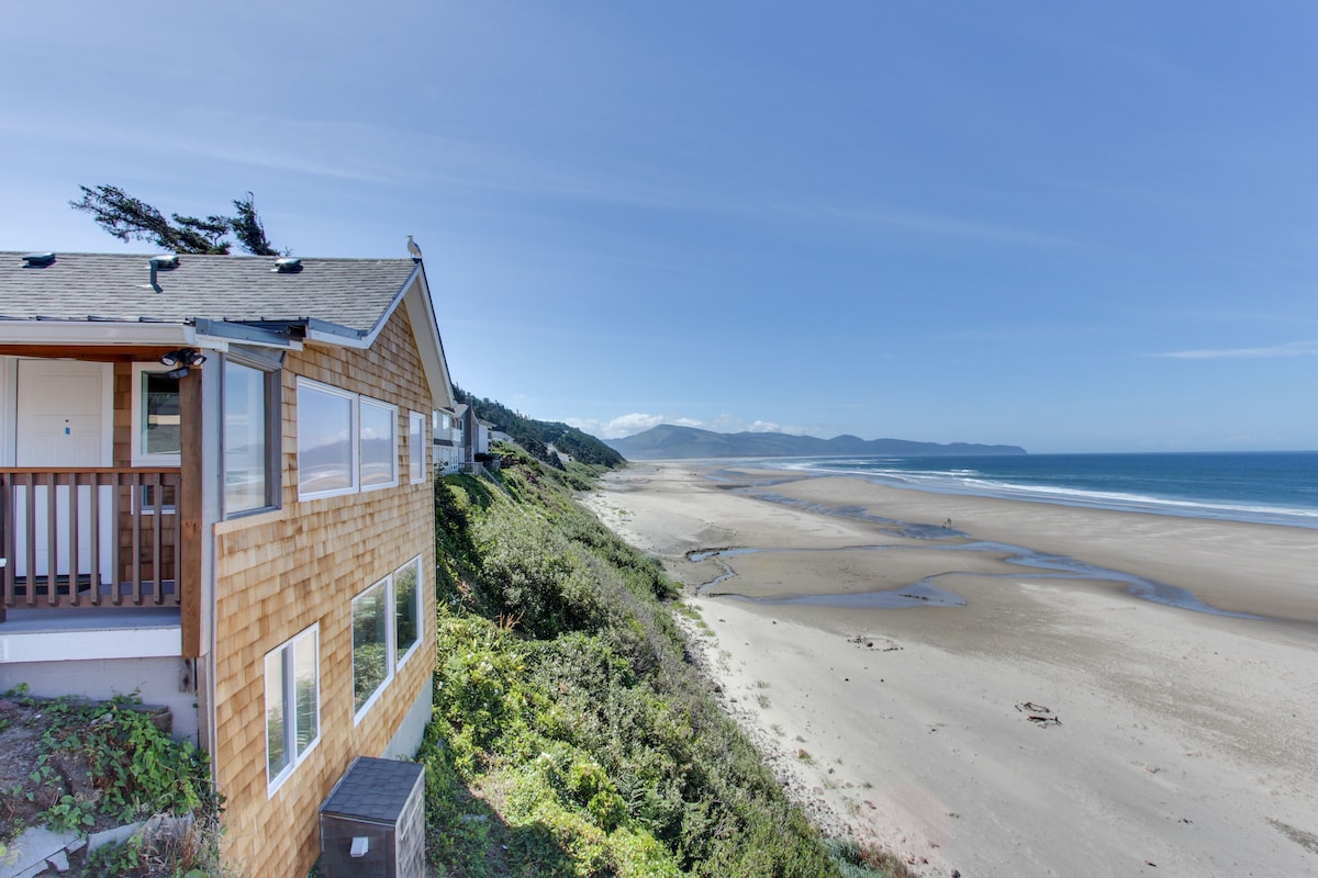 The image captures the exterior of a coastal inn with shingled siding, offering stunning views of a sandy beach and the Pacific Ocean. The horizon stretches toward rolling hills in the distance, under a clear, blue sky with minimal clouds.