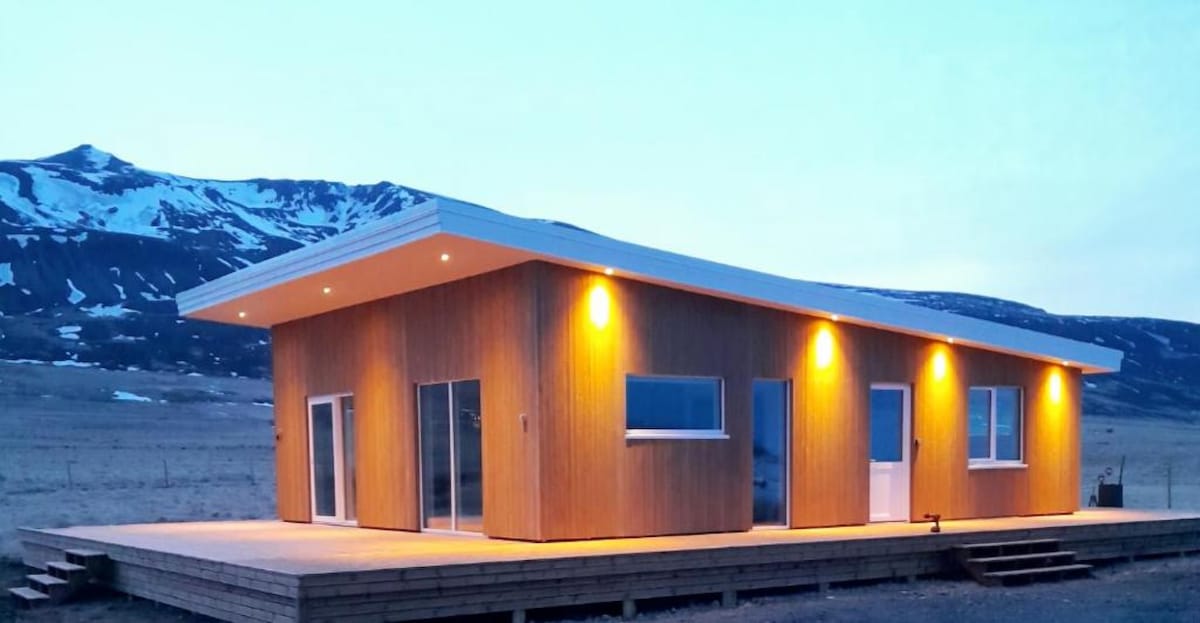 The modern house features a wooden exterior with large windows, allowing views of the surrounding landscape. Soft lighting illuminates the structure at dusk, while a spacious deck extends in front, providing a welcoming outdoor area. Snow-capped mountains are visible in the background.