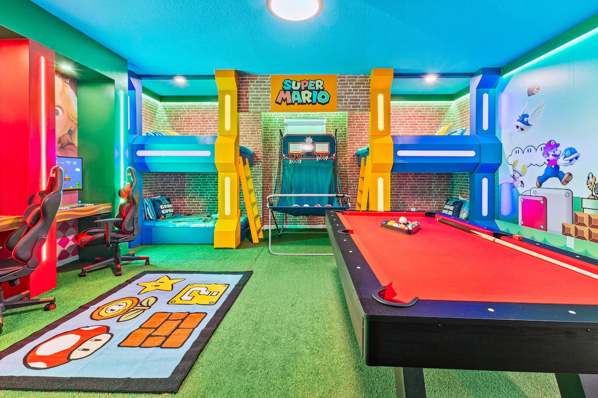A vibrant game room designed with a Super Mario theme features a red pool table and an arcade-style gaming station. Colorful wall graphics and playful carpet add to the inviting atmosphere. Comfortable gaming chairs are positioned near a desk, creating a fun environment for guests.