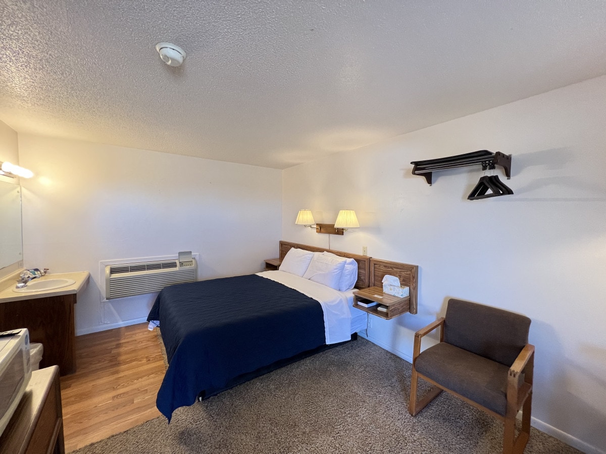 A spacious room features a large bed covered with a navy blue blanket. Two bedside lamps provide soft lighting. A chair and a small table are positioned next to the bed. A set of hangers is mounted on a wall shelf, while a bathroom sink is visible in the background.