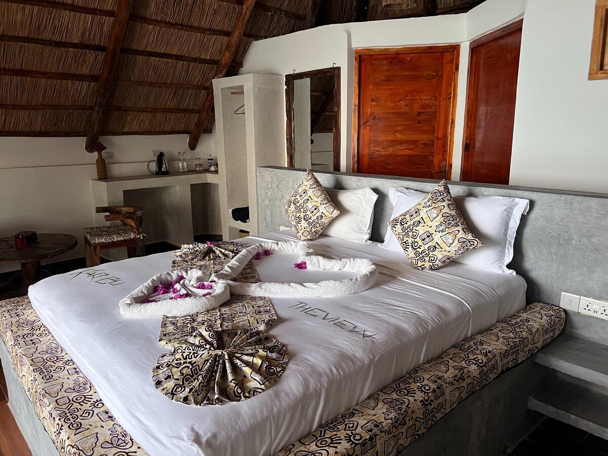 A cozy sleeping area features a spacious bed adorned with decorative pillows and floral accents. Light-colored linens are complemented by patterned bed covers. A shower area and a small kitchenette are visible in the background, alongside wooden doors and a thatched roof.