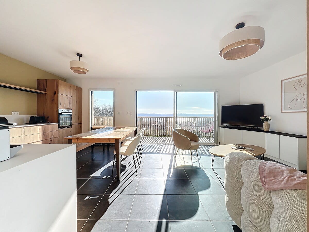 A modern living area features a large table surrounded by light-colored chairs. Natural light floods through expansive glass doors, revealing a view of the outdoors. A comfortable couch and a television are positioned in the corner, enhancing the open and airy feel of the space.