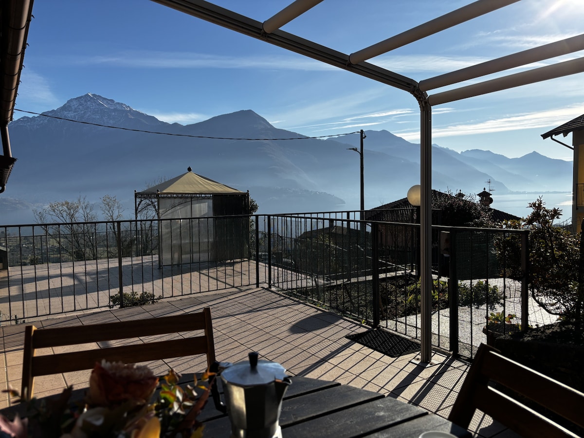 An expansive terrace provides a panoramic view of the surrounding mountains and valleys. The railing encloses the space, while sunlight casts soft shadows across the tiled flooring. A gazebo is visible in the distance, enhancing the serene outdoor environment.