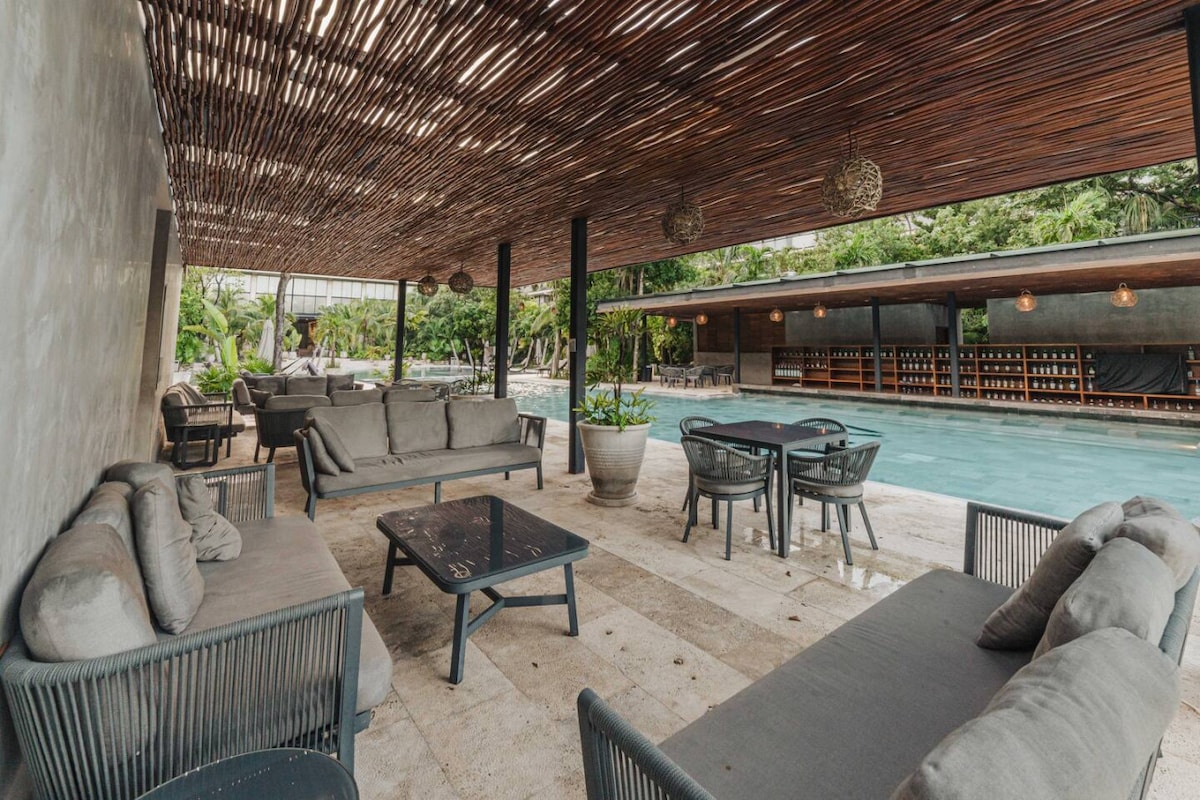 An outdoor lounge area features comfortable seating with grey cushions arranged around dark tables. Natural light filters through a wooden pergola, casting gentle shadows. The expansive pool is visible in the background, bordered by lush greenery, creating a serene setting for relaxation.
