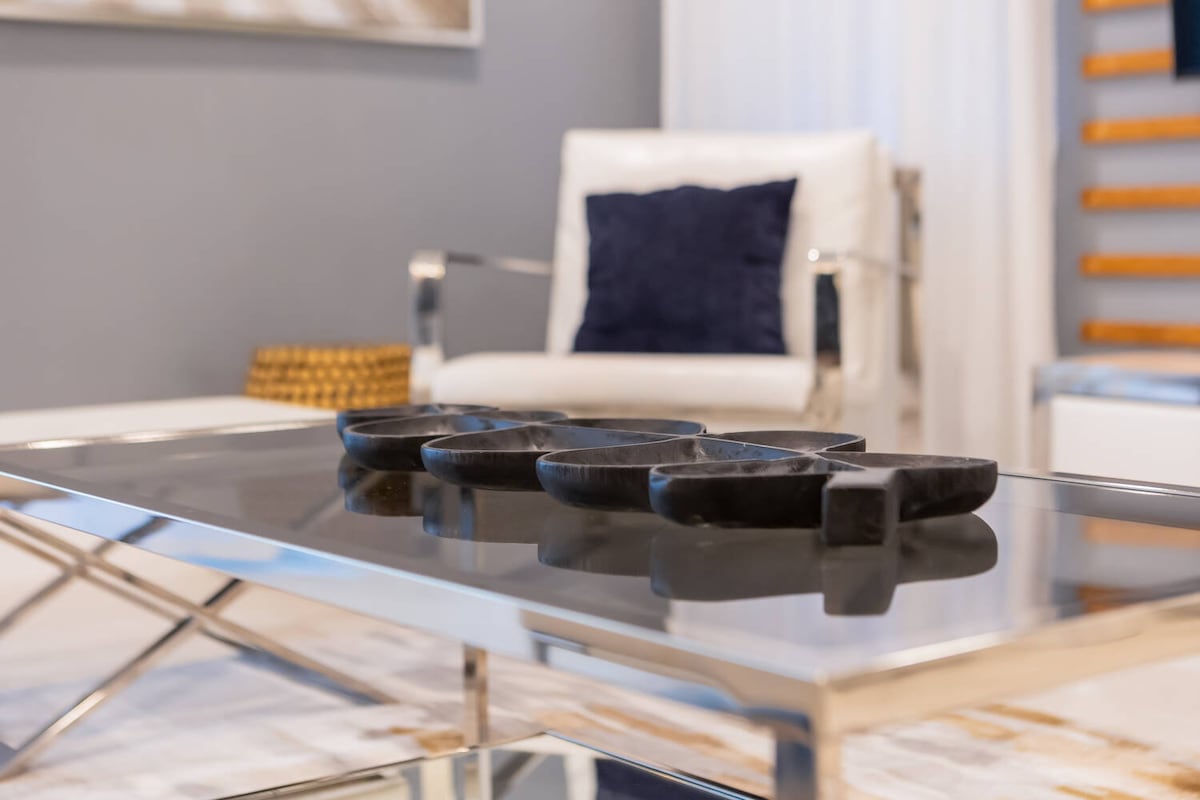 A sleek coffee table is highlighted with a reflective surface, displaying an arrangement of four black decorative bowls. A modern white armchair with a deep blue cushion is positioned beside the table, contributing to a sophisticated living area. Soft natural light filters through nearby sheer curtains.