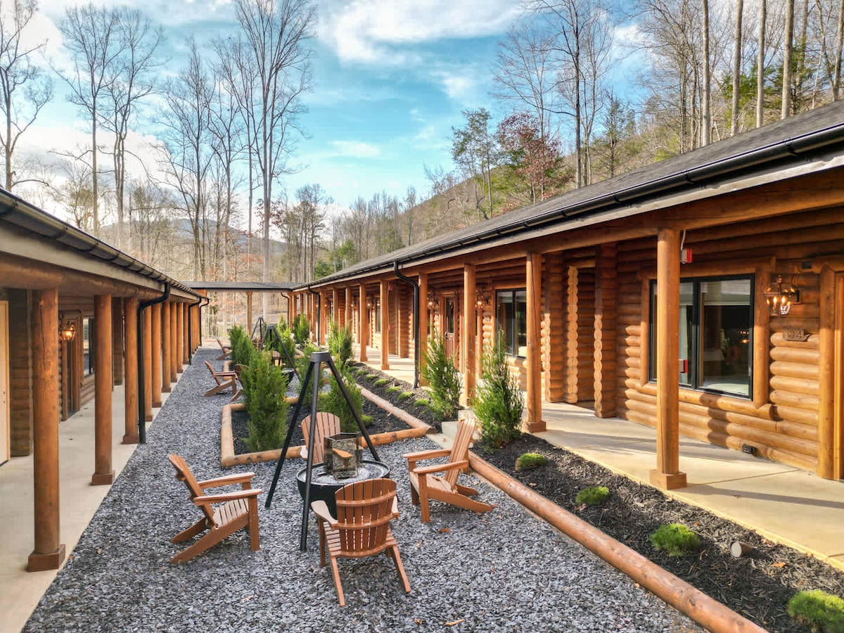 An inviting courtyard features rustic log cabins, surrounded by neatly arranged gravel pathways. Potted greenery adds natural elements to the space, alongside wooden chairs arranged around a fire pit, creating a welcoming spot for relaxation among the trees.