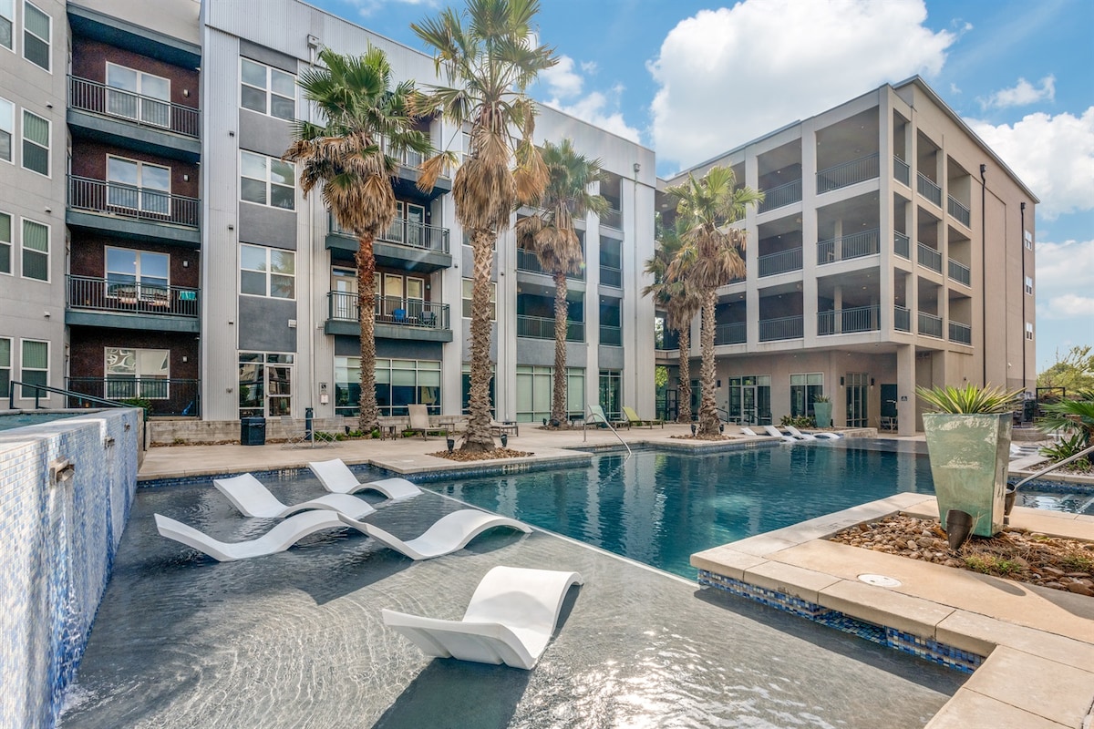 A resort-style swimming pool is surrounded by palm trees, featuring lounge chairs partially submerged in the water. The modern building facade stands in the background, showcasing large windows and balconies that enhance the inviting outdoor space.