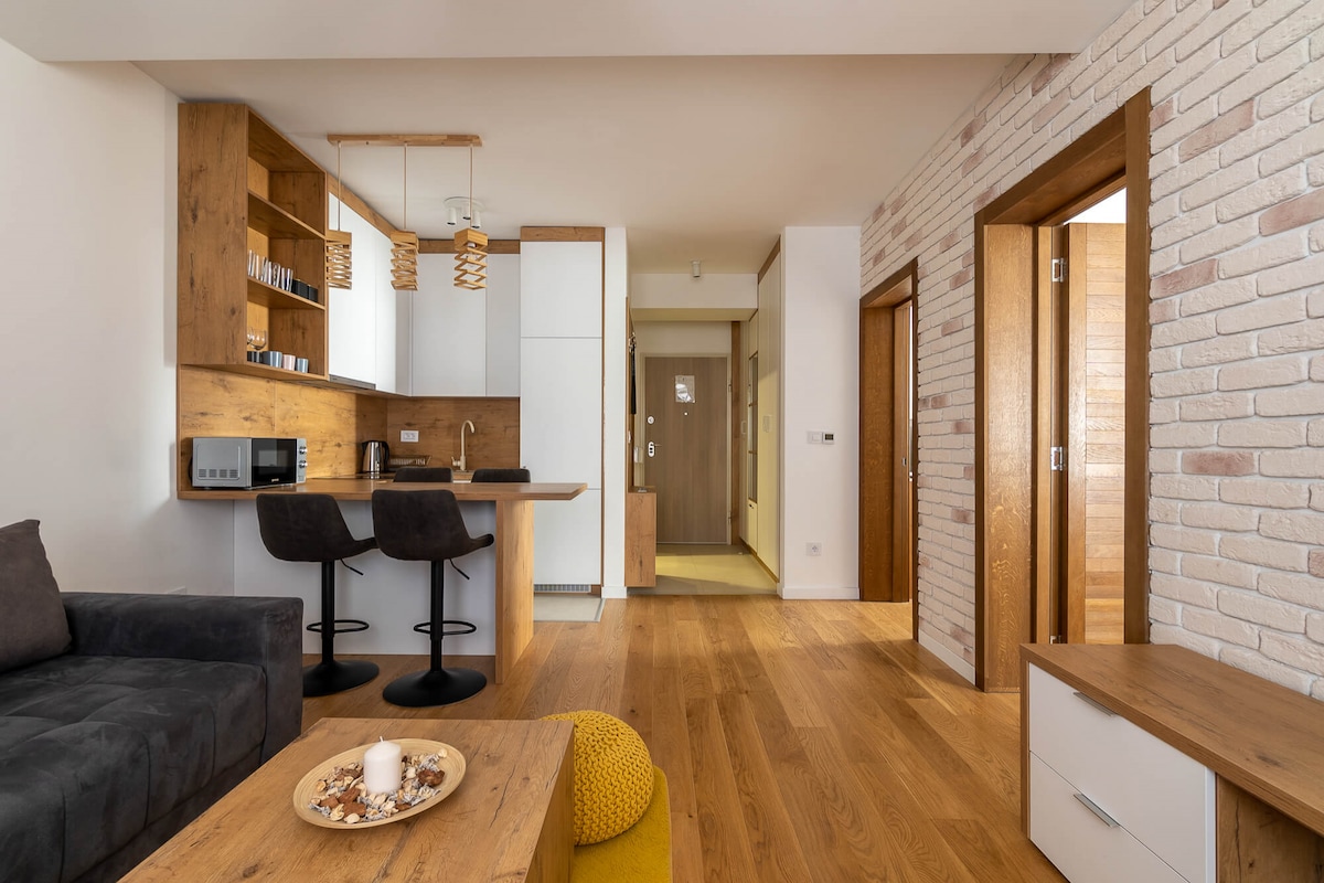An open living space is presented, featuring a comfortable gray sofa. A wooden dining table, complemented by two high stools, is positioned near a modern kitchenette. Light wooden flooring enhances the warm atmosphere, while recessed lighting and wall textures add visual interest.