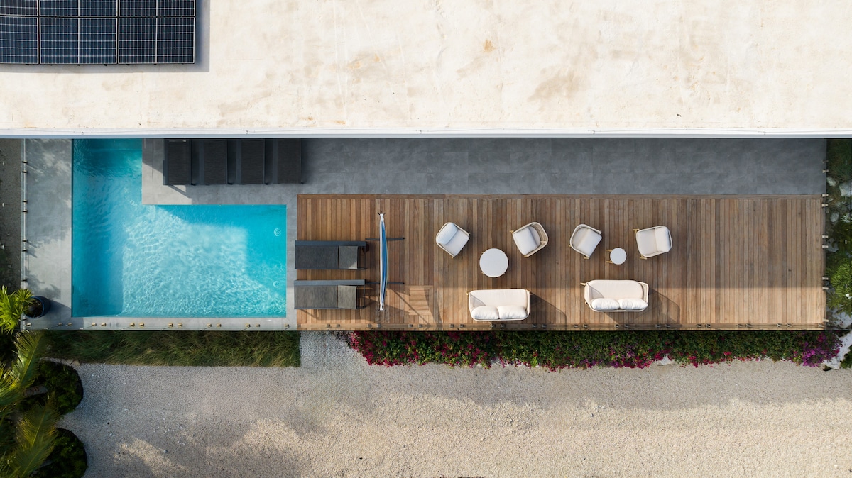 An overhead view showcases a sleek swimming pool adjacent to a wooden deck. The deck is furnished with a circular table and four lounge chairs, complemented by two comfortable sun loungers. Surrounding greenery and colorful flowers enhance the peaceful outdoor space.