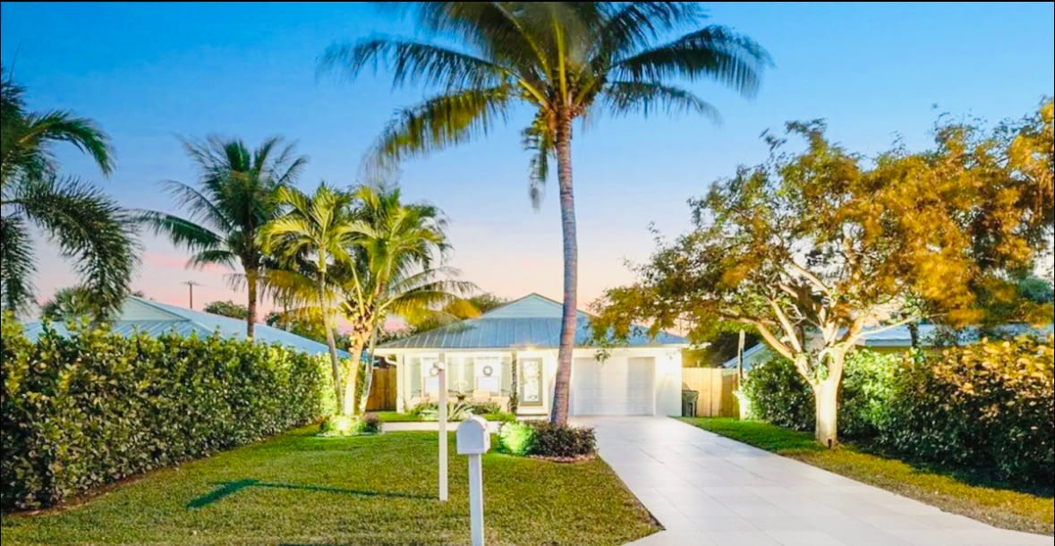 An inviting exterior of a house is surrounded by palm trees and lush greenery. A wide, paved pathway leads to the entrance, illuminated by soft landscape lighting. The sky transitions from daylight to dusk, enhancing the serene atmosphere of the property.