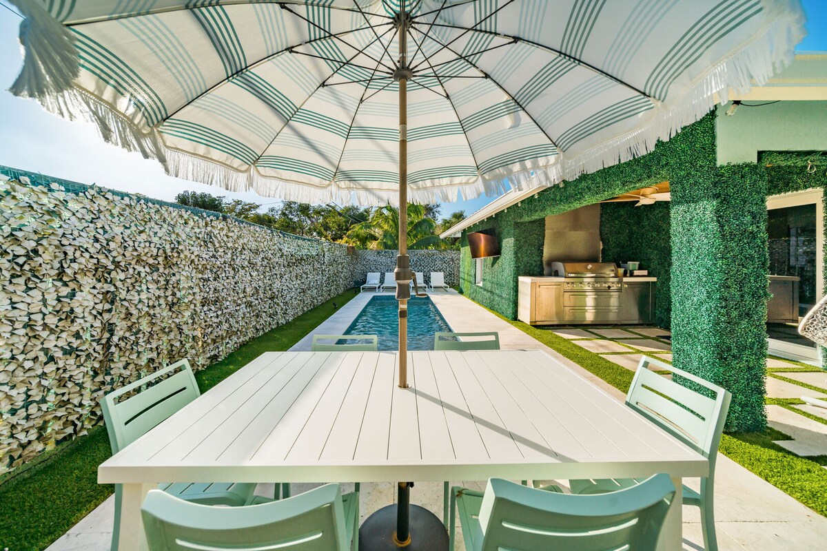 A dining table with four chairs is positioned beneath an oversized striped umbrella. In the background, a heated saltwater pool is framed by lush greenery and a stone-lined wall, providing a tranquil outdoor setting.