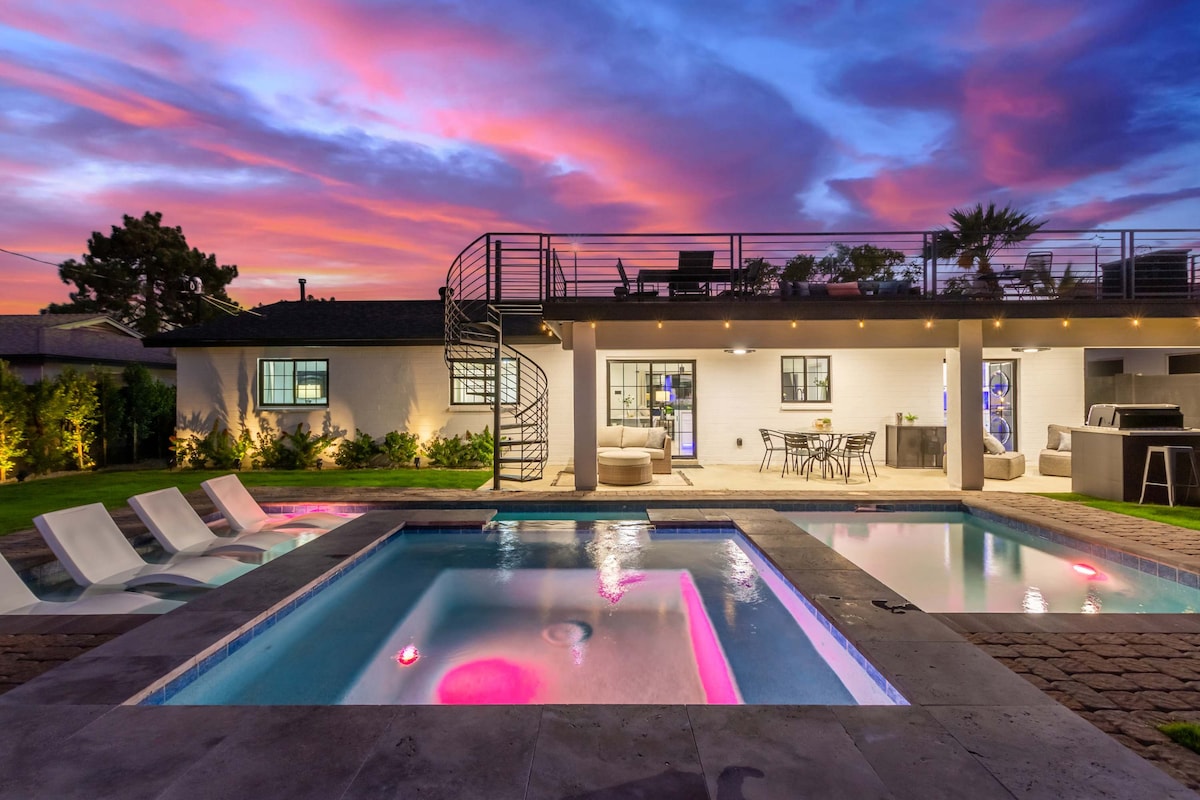 A modern outdoor space features a deep water pool beside an attached spa, with soft lighting enhancing vibrant colors. A covered patio offers seating and dining options. A spiral staircase leads to a rooftop terrace with additional seating, all set against a colorful evening sky.
