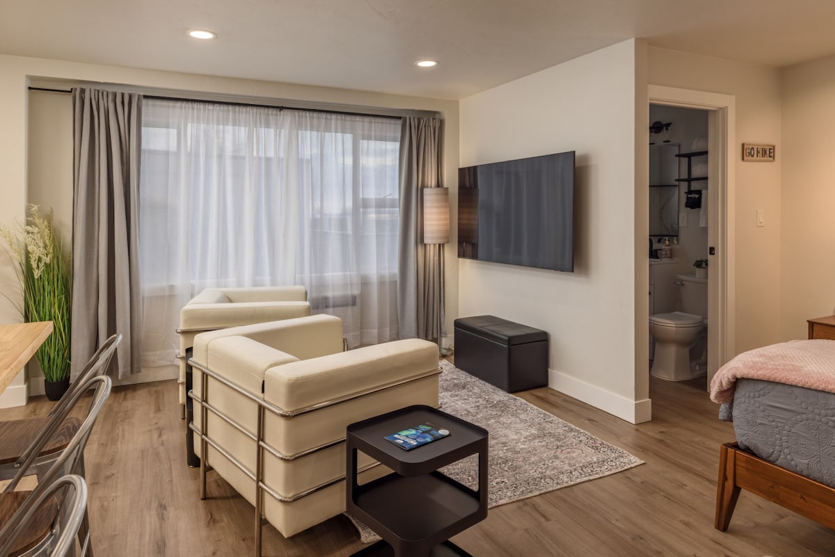 A cozy living area is furnished with two modern white armchairs and a sleek dark coffee table. A flat-screen TV is mounted on the wall, opposite a large window framed by neutral-toned curtains. The adjacent space leads to an open kitchenette and a bathroom.
