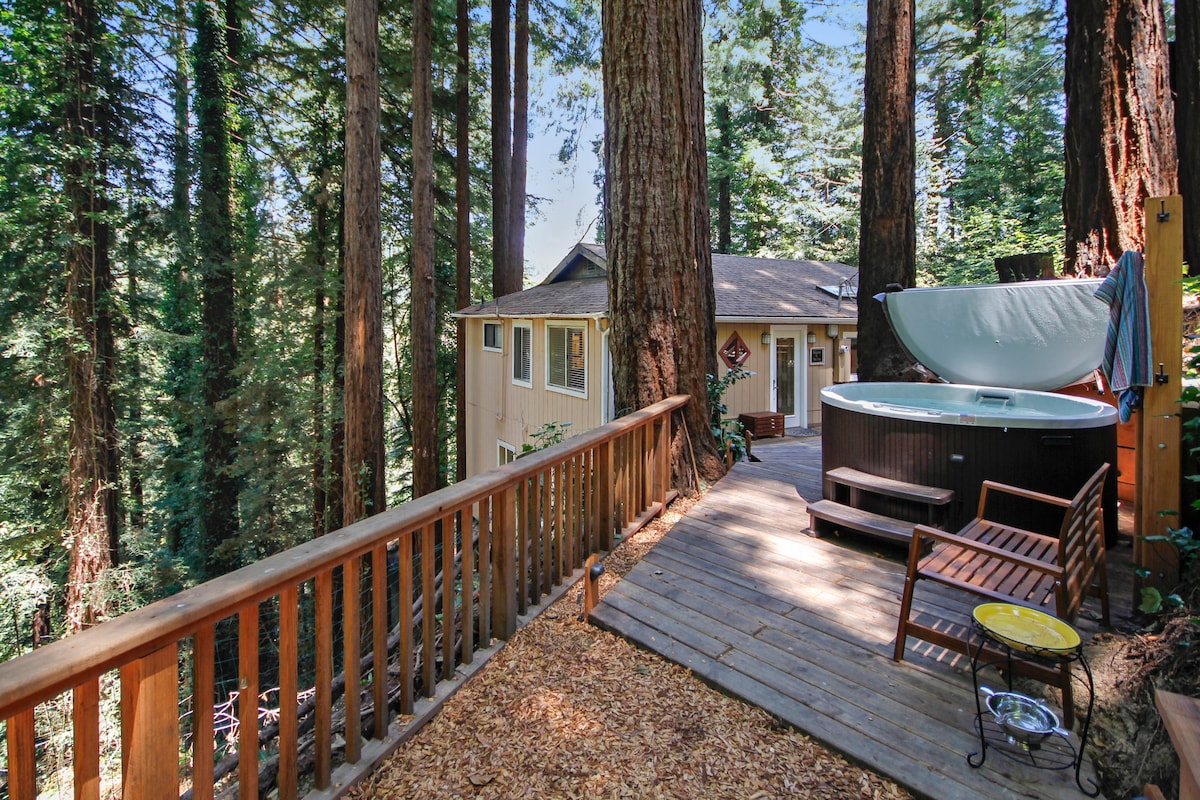 A wooden deck is surrounded by towering redwood trees, creating a secluded setting. A hot tub sits to one side, with a cover partially opened, inviting relaxation. A bench and a small table are positioned nearby, enhancing the outdoor space's functionality.