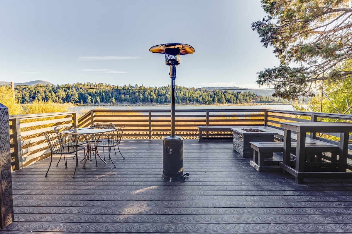 An expansive deck features a circular dining table with four chairs, allowing for outdoor meals. A patio heater stands tall, providing warmth during cooler evenings. A fire pit surrounded by seating completes this inviting space, all set against a backdrop of serene lake views.