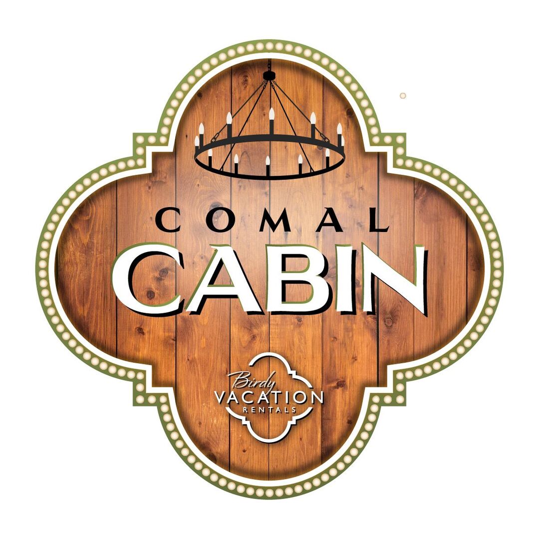 A decorative sign displays the name 'Comal Cabin' prominently against a wooden background. The design features an elegant border and a chandelier motif above the text, which indicates the cabin is part of Birdy Vacation Rentals.