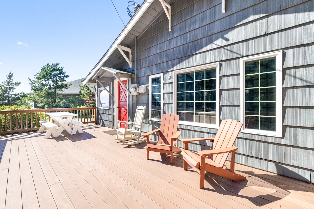 A spacious outdoor deck features two wooden adirondack chairs and a dining table, providing ample seating for relaxation and dining. Large windows in the cabin allow natural light to enhance the inviting atmosphere. The backdrop includes trees, adding a touch of nature to the scene.