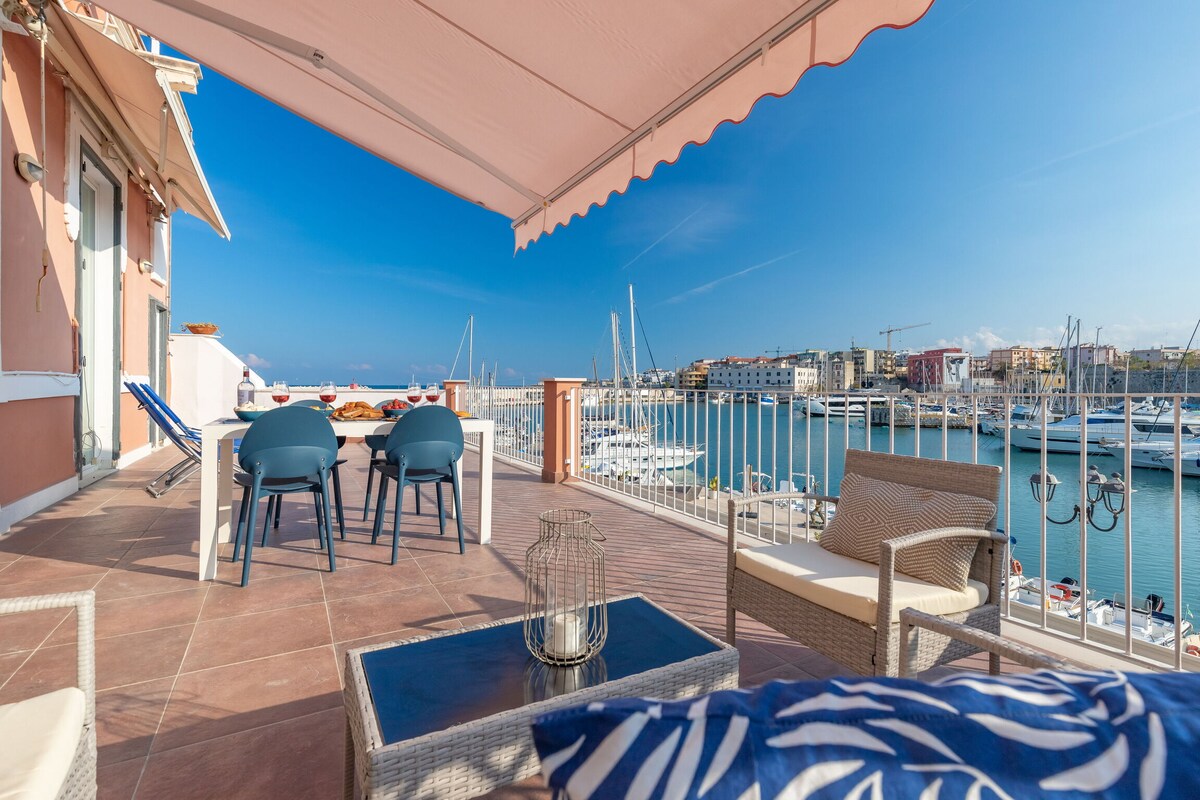 A spacious terrace features comfortable seating areas with armchairs and a dining set, overlooking the marina. A view of the tranquil water and moored boats is complemented by a clear blue sky, creating an inviting outdoor space for relaxation and meals.