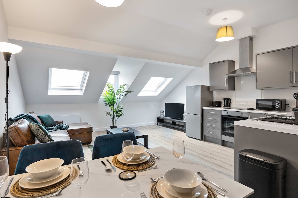 A bright, modern kitchen and dining area are shown, featuring a sleek gray kitchen with stainless steel appliances. A comfortable seating area is visible in the background, complemented by two skylights offering natural light. The dining table is elegantly set, ready for meals.